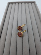 Runda örhängen från Nayuga Jewels med jordgubbskvarts infattad i guldpläterat stål, placerade på en grå smyckesbricka.