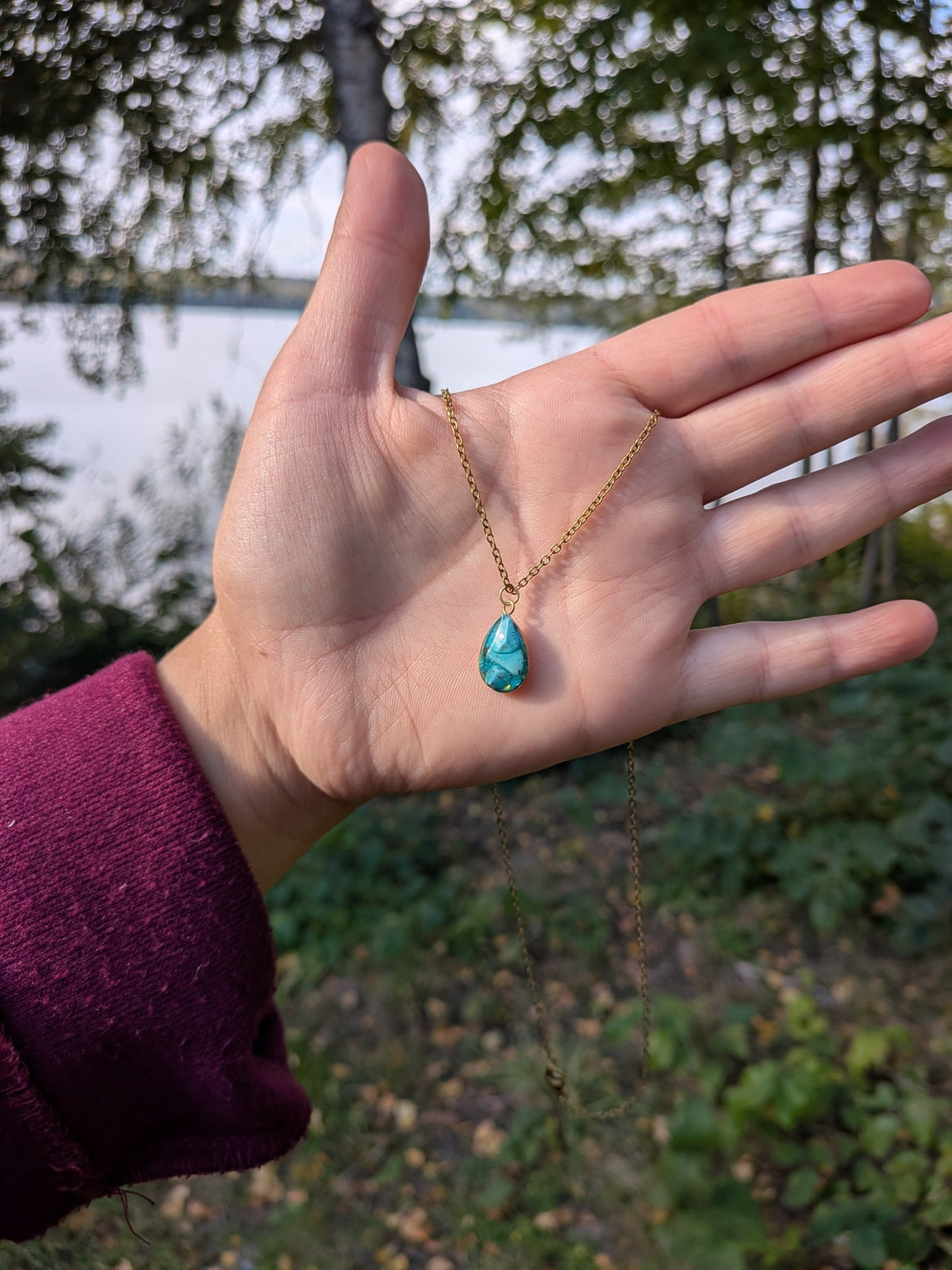 Citrinmagi droppformat halsband med turkos sten i guldfärgad infattning och tunn kedja som hålls i en hand.