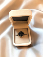 Citrinmagi ring i guld med rund infattning fylld av svarta glittrande stenfragment i en beige smyckesask.