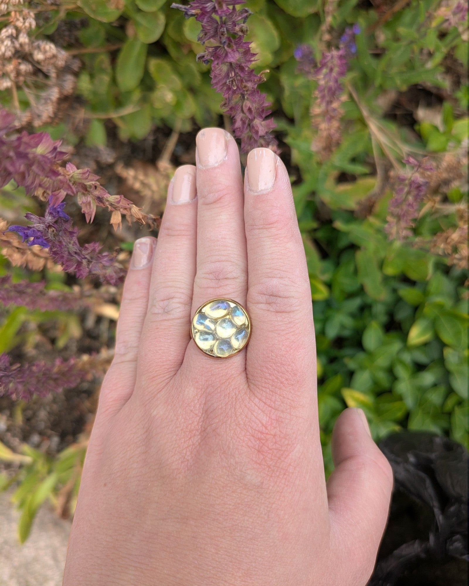 Citrinmagi handgjord guldring med rund skimrande sten på ett finger mot en bakgrund av lila blommor.