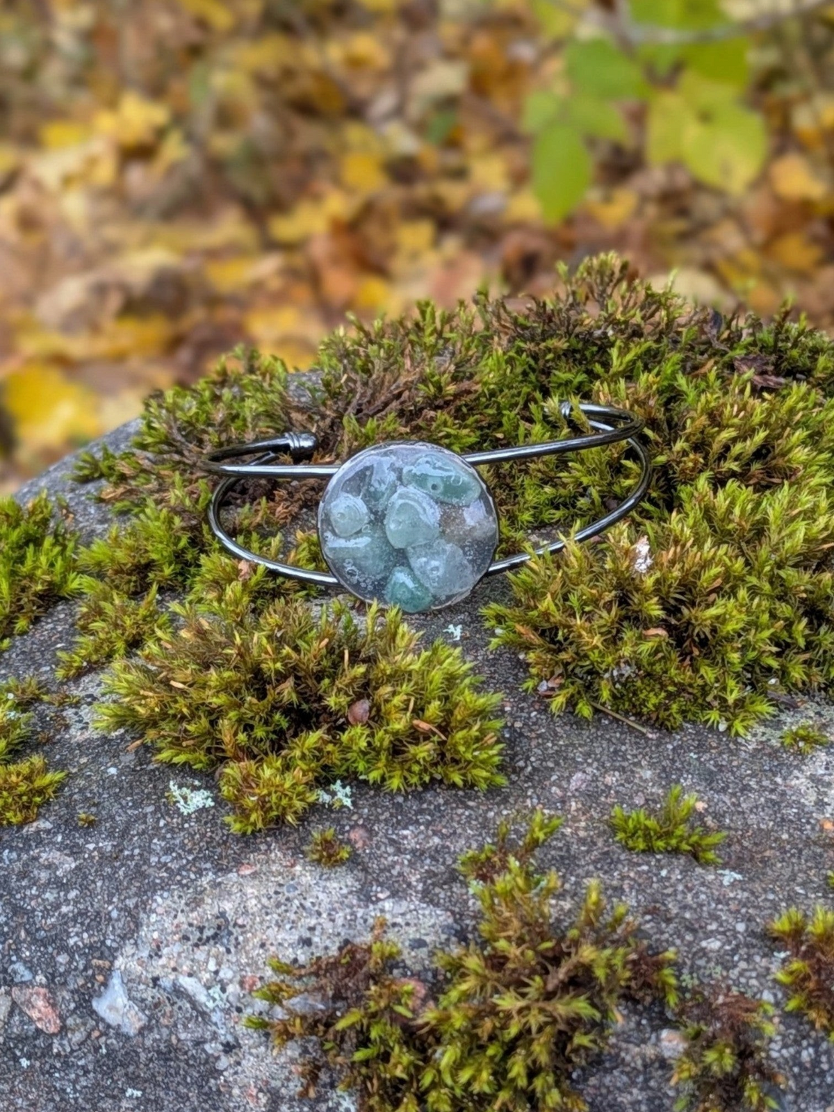 Citrinmagi handgjort cuff-armband i mörk metall med gröna slipade ädelstenar placerat på en mossa-täckt sten.