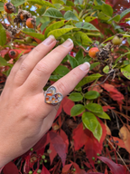 Citrinmagi hjärtformad ring i silverfärgad metall med mosaik av bärnstensliknande stenar på ett finger i en trädgård.