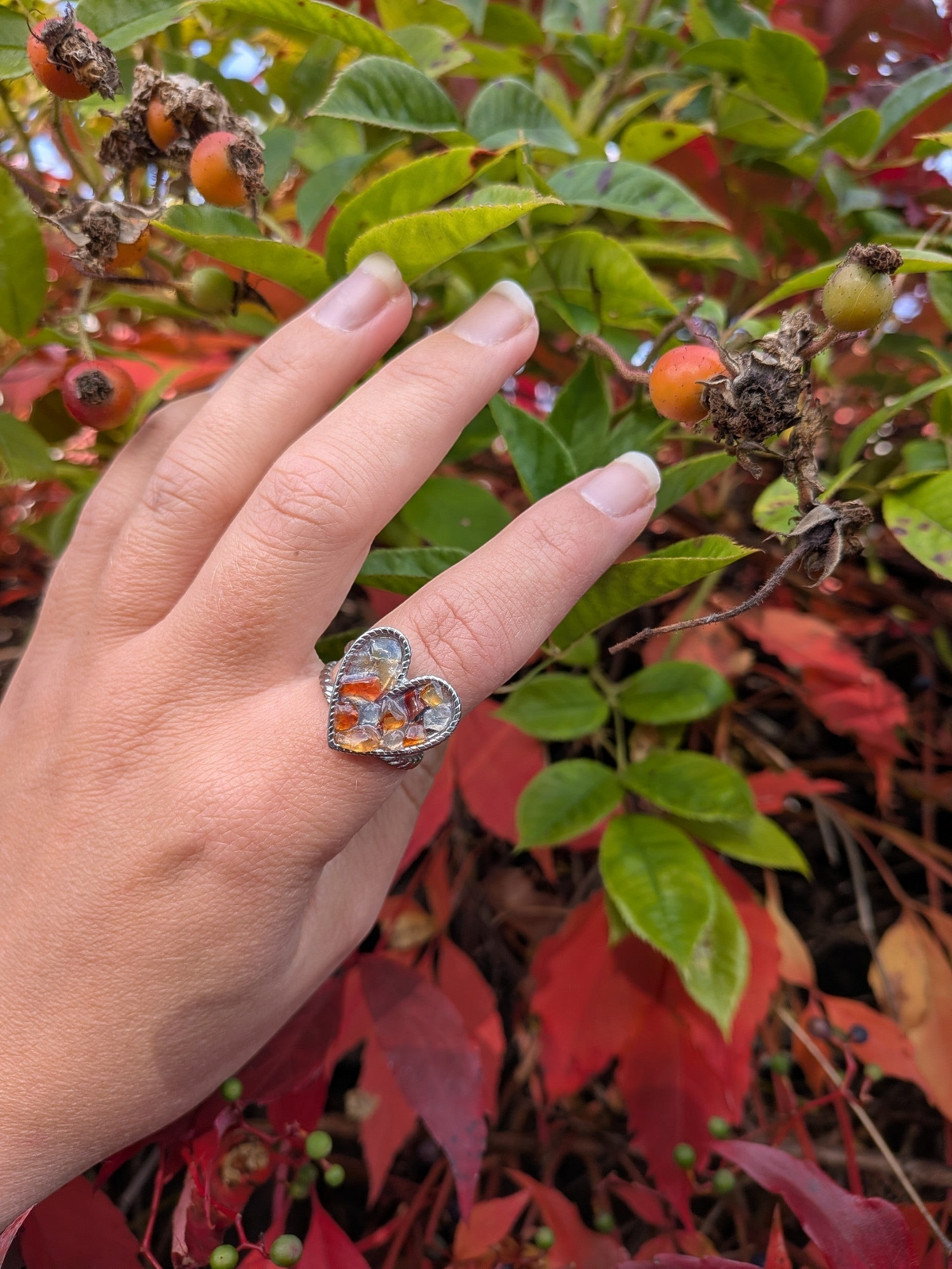 Citrinmagi hjärtformad ring i silverfärgad metall med mosaik av bärnstensliknande stenar på ett finger i en trädgård.