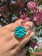 Citrinmagi ring i guld med turkosa howlit-kristaller, hållen mot en bakgrund av blommande rosa grenar.