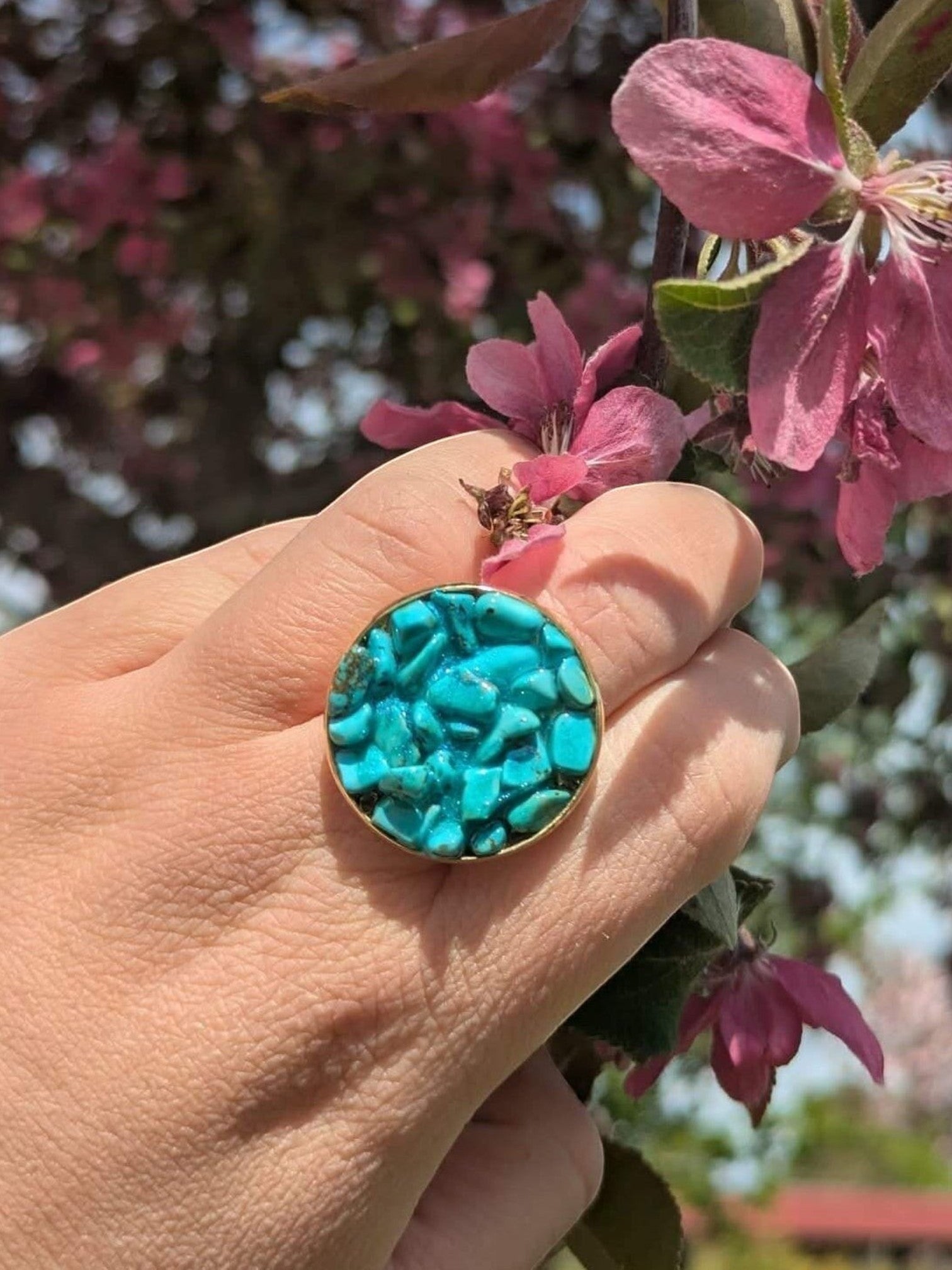 Citrinmagi ring i guld med turkosa howlit-kristaller, hållen mot en bakgrund av blommande rosa grenar.