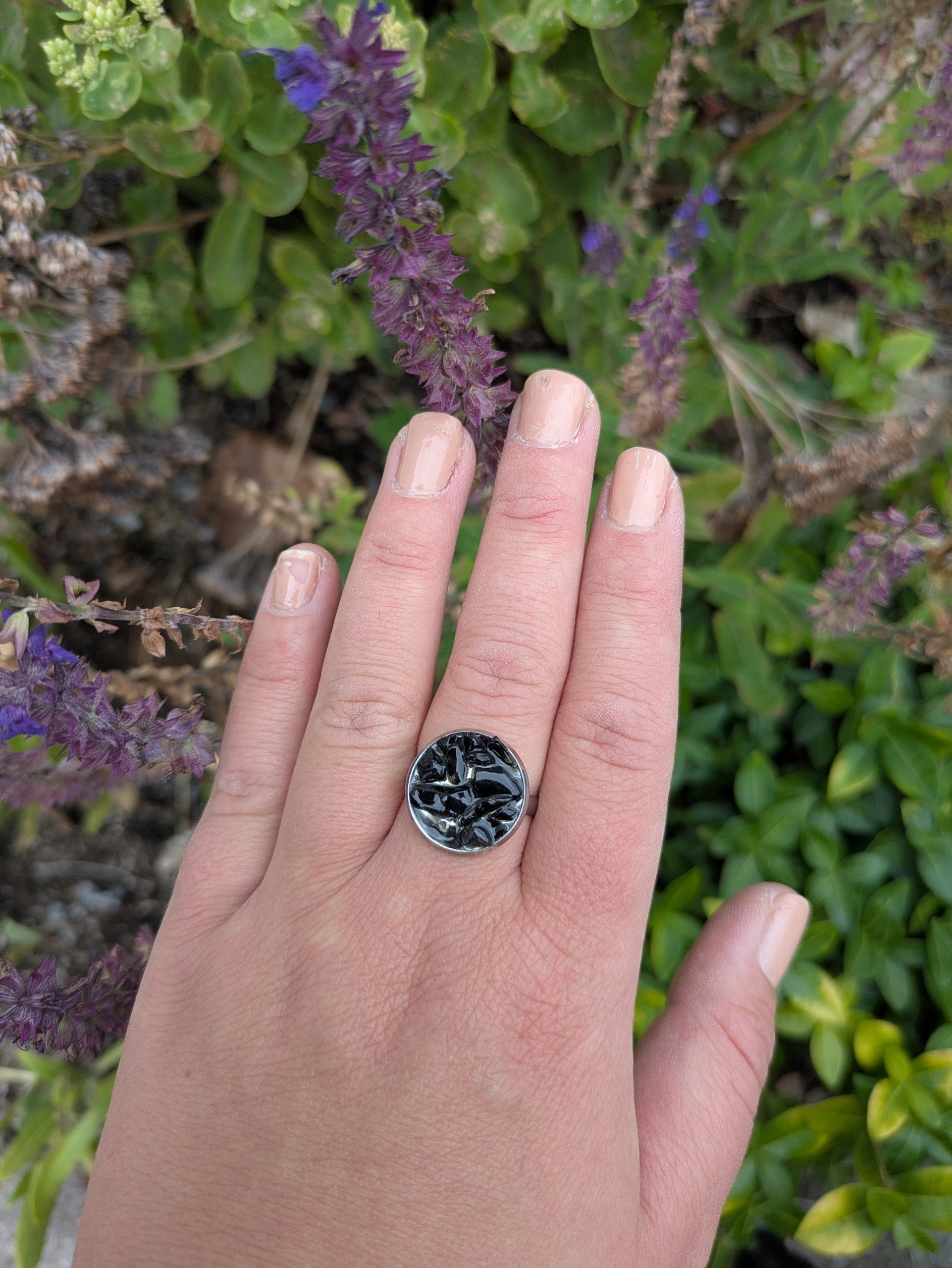Citrinmagi ring med rund infattning och svart texturerad yta på en hand mot en bakgrund av lila blommor.