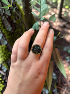 Citrinmagi rund guldring med glittrande svarta stenfragment buren på ett finger mot en bakgrund av blommor